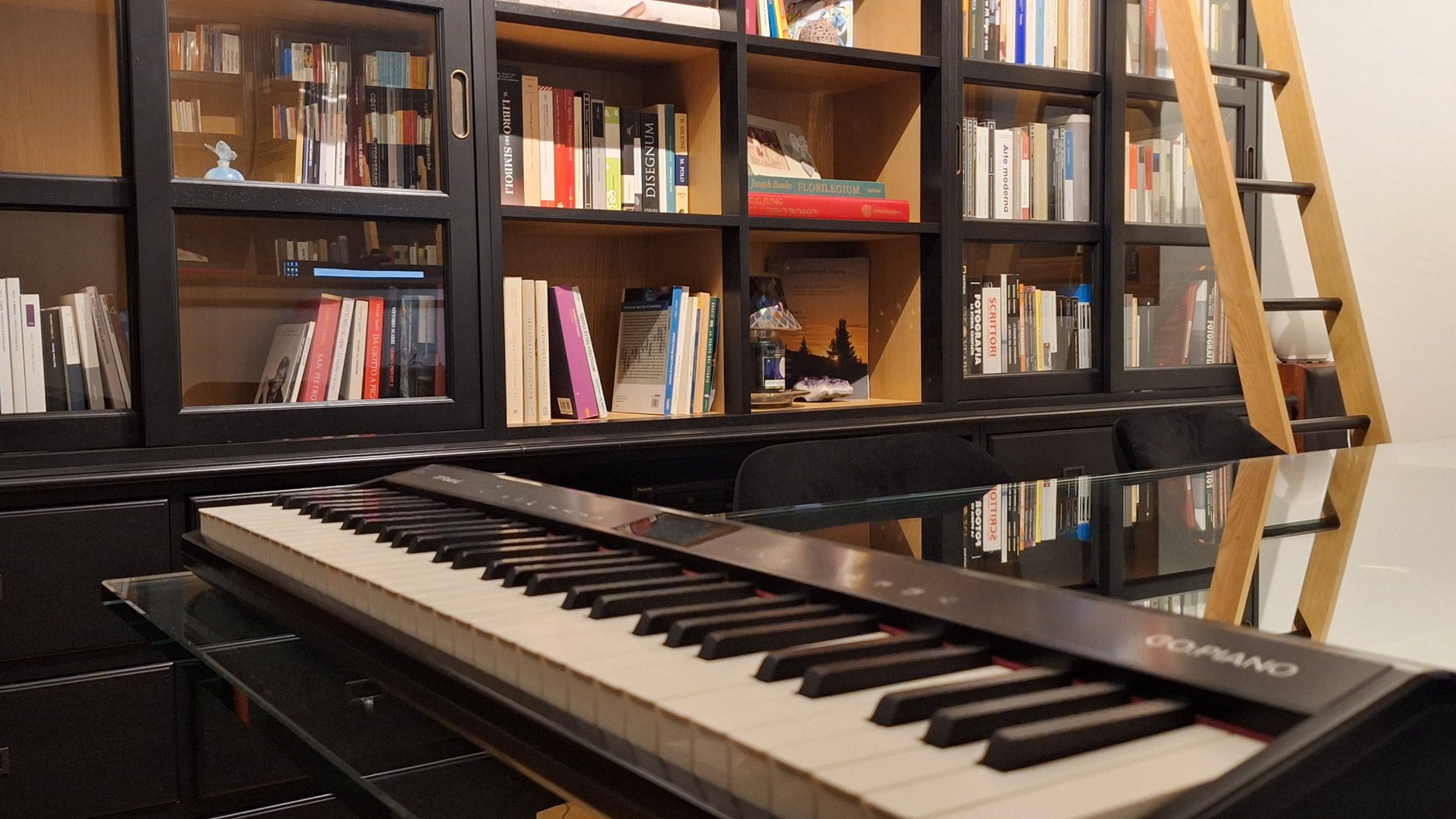 Lo studio di Riccardo Donati: un pianoforte digitale su una scrivania di vetro, davanti a una libreria nera piena di libri, con una scala di legno.