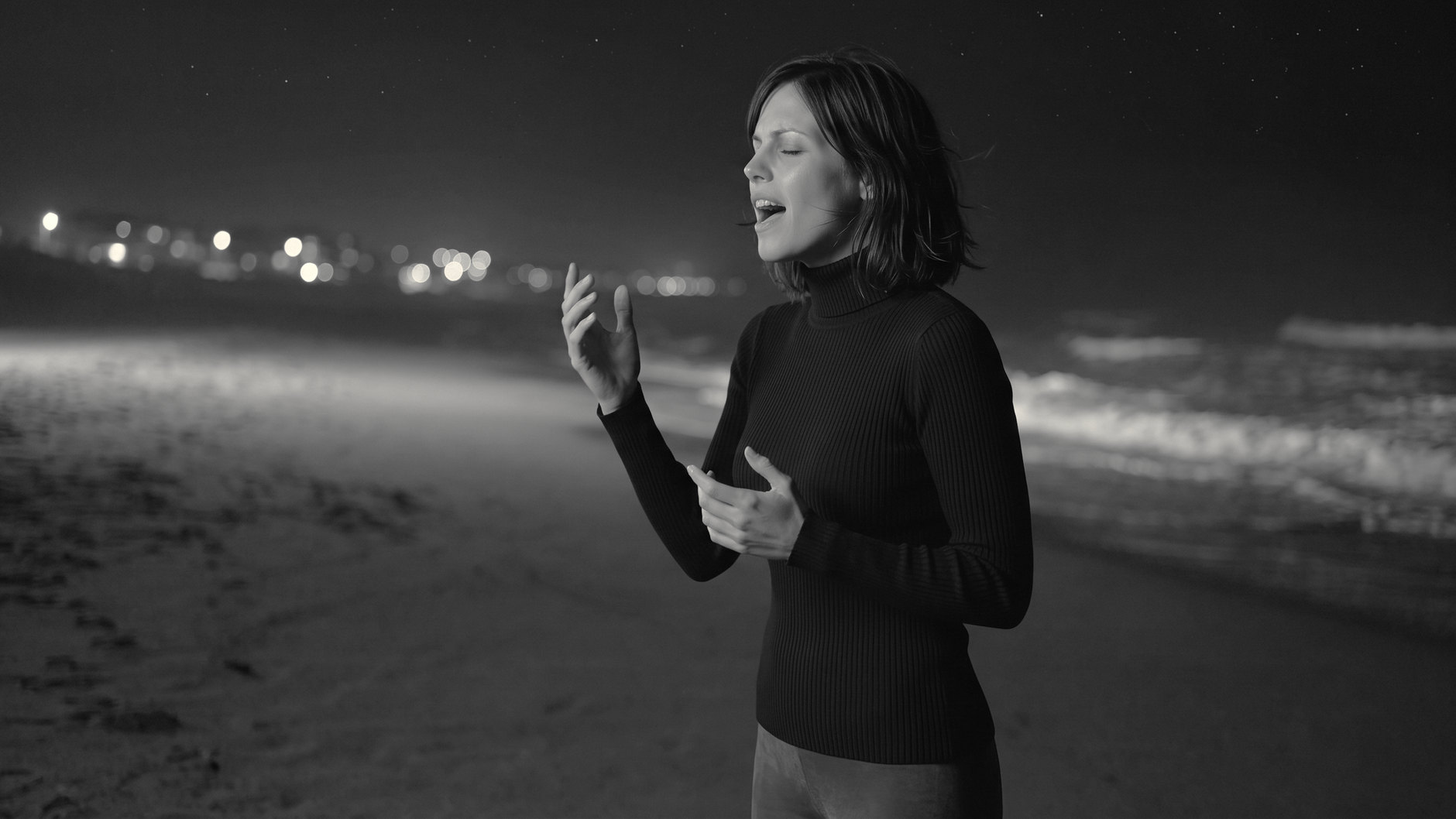 Alma canta sulla riva di notte, occhi chiusi, una mano sollevata; le luci del paese sfocate alle spalle. Immagine in bianco e nero.