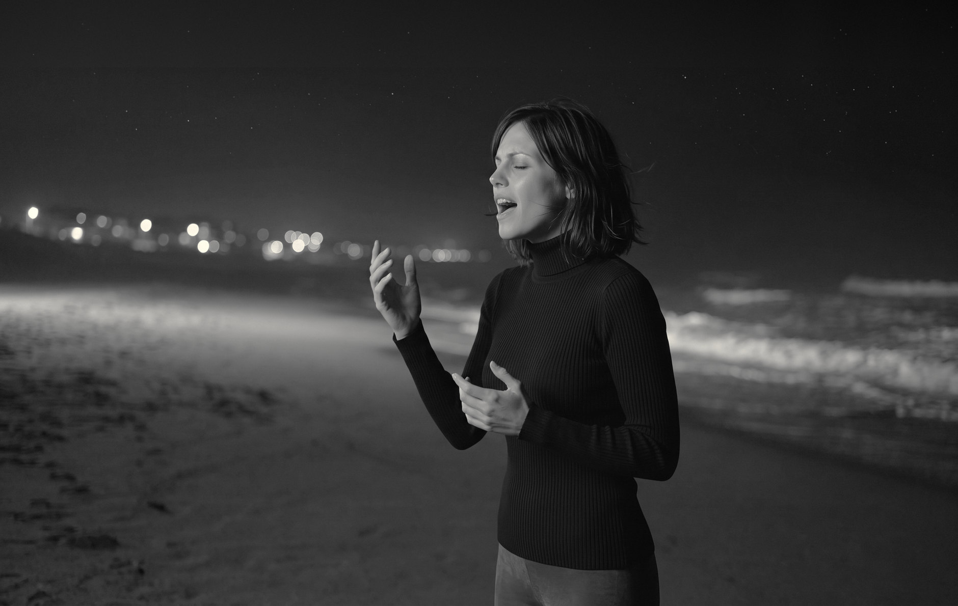 Alma canta sulla riva di notte, occhi chiusi, una mano sollevata; le luci del paese sfocate alle spalle. Immagine in bianco e nero.